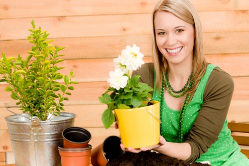 A woman with blonde hair and a cheerful expression sits outdoors on a wooden deck or patio area, holding a bright yellow flower pot containing green leafy plants with white flowers. In front of her, there are various gardening pots and containers, including a metallic bucket with a lush green shrub, a black plastic pot, and an unglazed terracotta pot. The background features a warm-toned wooden fence or wall, indicating a well-maintained garden or backyard space suitable for gardening and outdoor plant care. The scene is well-lit with natural daylight, highlighting the fresh, vibrant colours of the plants and the woman's green apron and layered necklaces. This setting exemplifies a typical outdoor gardening or lawn maintenance environment that Lawn Mowing Osterley might service, with emphasis on healthy plant growth and garden organisation, situated within the Osterley or surrounding West London area.